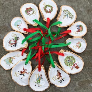 May include: A collection of twelve oyster shell ornaments, each with a gold-painted edge and a unique hand-painted illustration. The ornaments feature festive designs, numbers, and red and green ribbons for hanging. A decorative holiday theme.