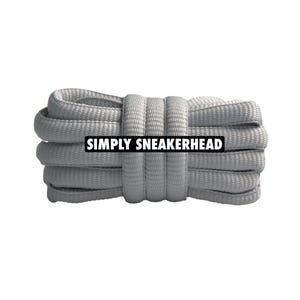 Peut inclure: Un paquet de lacets gris clair, enroulés serrés et fixés au milieu. Le texte "SIMPLY SNEAKERHEAD" est affiché dans un rectangle noir sur les lacets.