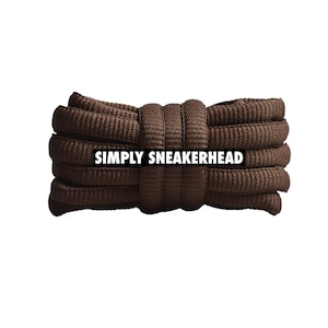 Può includere: Un fascio di lacci marroni, strettamente arrotolati e disposti orizzontalmente. I lacci sono di un colore marrone scuro uniforme con una superficie strutturata. Il testo "SIMPLY SNEAKERHEAD" è stampato in bianco al centro dei lacci.