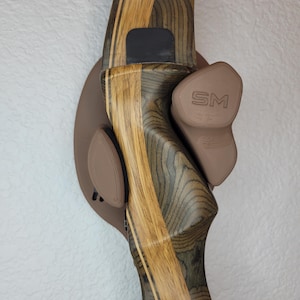 May include: A wooden bow with a black and green wood grain pattern is mounted on a brown wall mount with two brown rubber pads. The wall mount has the letters "SM" printed on it.