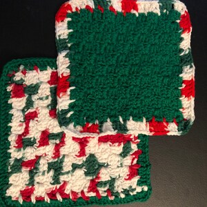 May include: Two green crocheted dishcloths with red and white trim. The dishcloths are made with a textured stitch.
