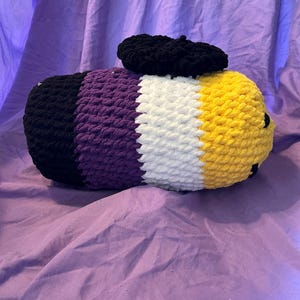 May include: A handmade, plush, non-binary pride flag pillow. The pillow is a cylinder shape with black, purple, white, and yellow stripes. A small, black, beret-style hat sits on top of the pillow. The pillow is on a purple surface.