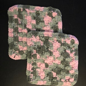 May include: Two crocheted dishcloths in a pink and gray color pattern. The dishcloths are square shaped and have a textured surface.