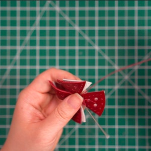 Peut inclure: Une fleur en papier rouge et blanche avec un cordon rouge attach&eacute;. La fleur est tenue par une main sur un tapis de coupe vert.