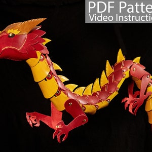 Op de afbeelding: Een gelede drakenpop van leer, in rood en geel. De draak heeft een lange staart, vier poten en een kop met hoorns. De draak staat in een vliegende houding.
