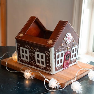 May include: A brown leather gingerbread house-shaped container with white stitching and accents. The house has white window frames, a red door, and a candy cane design. It sits on a wooden board with string lights.