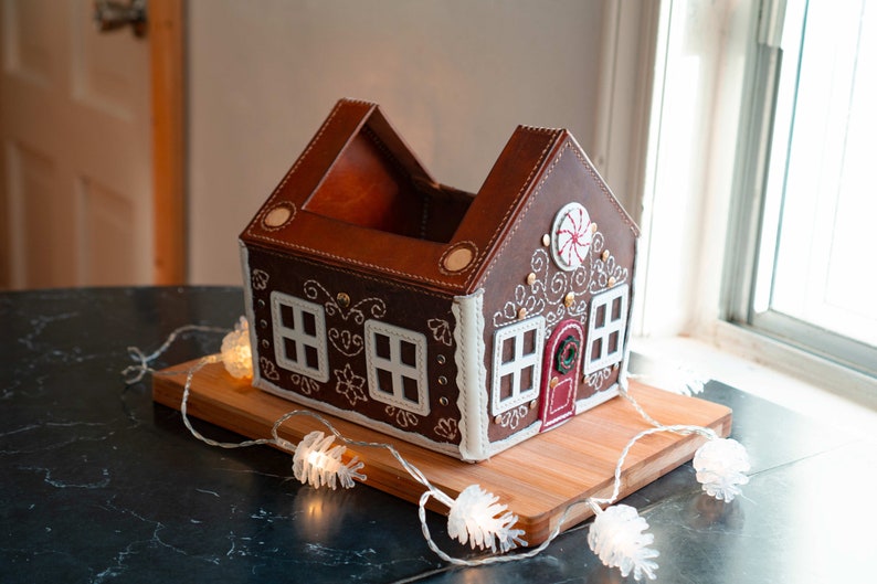 May include: A brown leather gingerbread house-shaped container with white stitching and accents. The house has white window frames, a red door, and a candy cane design. It sits on a wooden board with string lights.