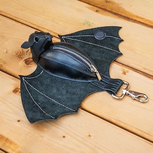 May include: A black leather bat-shaped fanny pack with a zippered pouch and silver-tone hardware. The wings are made of dark green suede and stitched with white thread. The bag is on a wooden surface.