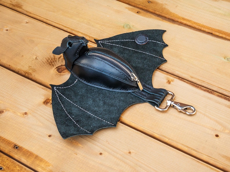 May include: A black leather bat-shaped fanny pack with a zippered pouch and silver-tone hardware. The wings are made of dark green suede and stitched with white thread. The bag is on a wooden surface.