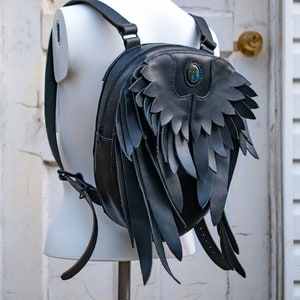 May include: A black leather backpack with wings. The backpack has a round shape and a blue gemstone accent.