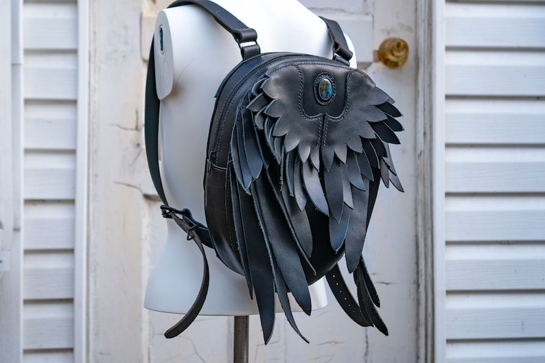 May include: A black leather backpack with wings. The backpack has a round shape and a blue gemstone accent.