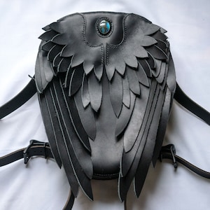 May include: Black leather backpack with wings and a blue gemstone accent. The wings are made of layered leather and are detailed with stitching.