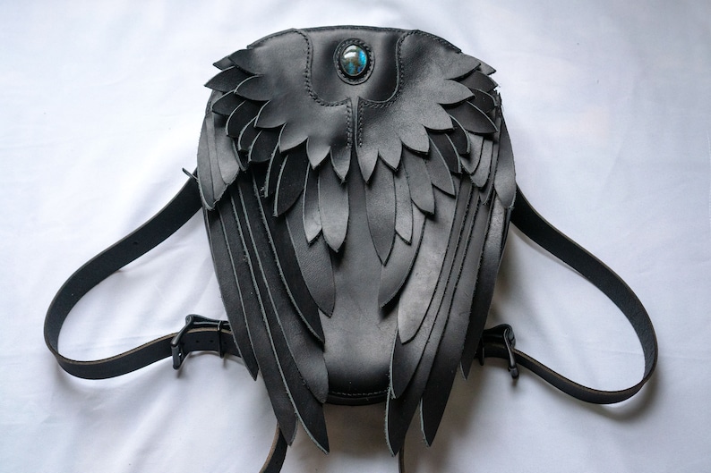 May include: Black leather backpack with wings and a blue gemstone accent. The wings are made of layered leather and are detailed with stitching.