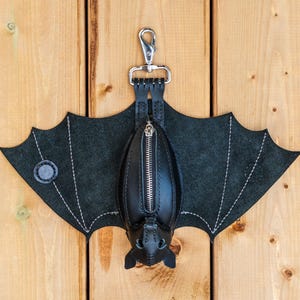 May include: A black leather bat-shaped keychain wallet with a silver zipper and a metal clasp. The wings are made of dark gray suede with white stitching. The wallet is hanging on a wooden surface.