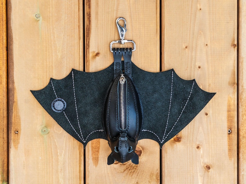 May include: A black leather bat-shaped keychain wallet with a silver zipper and a metal clasp. The wings are made of dark gray suede with white stitching. The wallet is hanging on a wooden surface.