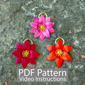 Può includere: Tre ornamenti floreali in pelle fatti a mano nei toni del rosa, rosso e arancione, ciascuno con un anello dorato e un centro giallo. I fiori sono disposti su una superficie grigia strutturata. Il testo in basso recita "PDF Pattern Video Instructions."