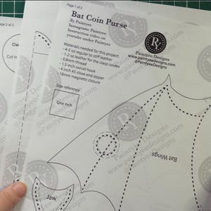 May include: Three white paper sheets with printed instructions and patterns for a bat-shaped coin purse. The sheets include text, dotted lines, and a size reference of one inch. The sheets are held over a green grid.