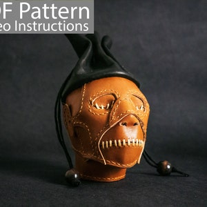 May include: A brown leather mask with stitched details, resembling a human face. The mask is attached to a black leather hat with a long cord.