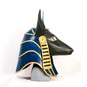 May include: A blue and gold Anubis mask with a black leather face and gold accents. The mask is on a gray stand.