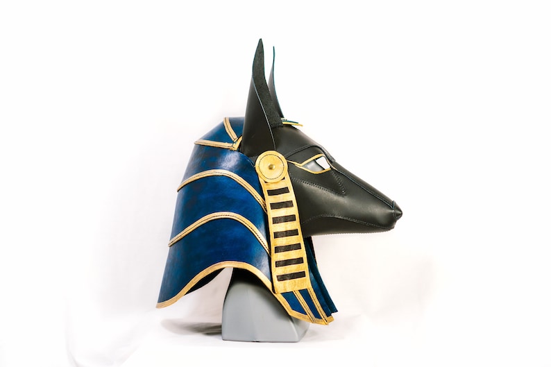 May include: A blue and gold Anubis mask with a black leather face and gold accents. The mask is on a gray stand.