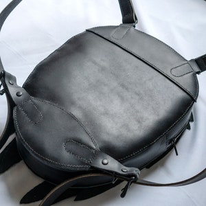 May include: A black leather backpack with adjustable straps and a zipper closure. The backpack has a round shape and a flap with a buckle closure.
