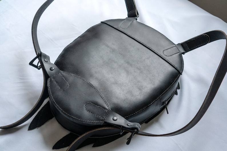 May include: A black leather backpack with adjustable straps and a zipper closure. The backpack has a round shape and a flap with a buckle closure.