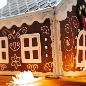 May include: A brown gingerbread house with white trim and decorative stitching. The house features white-framed windows, a decorative roof, and a small wreath. The house is decorated with small silver studs and sits on a wooden surface.