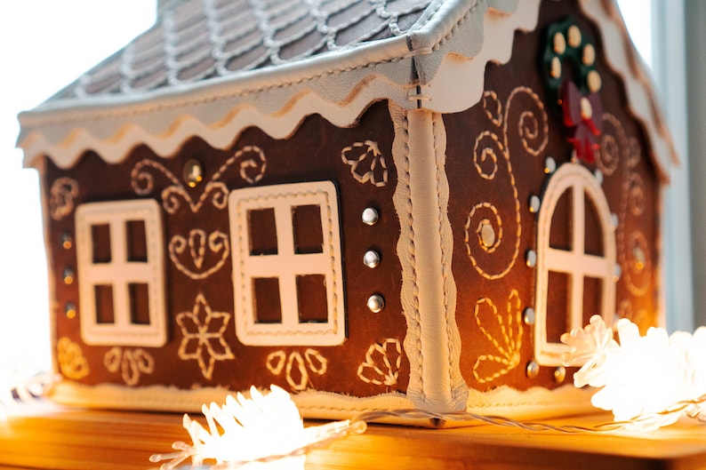 May include: A brown gingerbread house with white trim and decorative stitching. The house features white-framed windows, a decorative roof, and a small wreath. The house is decorated with small silver studs and sits on a wooden surface.