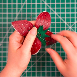 May include: A red leather strawberry with white dots and a green leather leaf is being held by two hands. The strawberry is on a green cutting mat.
