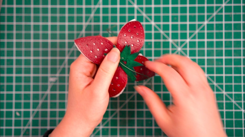 May include: A red leather strawberry with white dots and a green leather leaf is being held by two hands. The strawberry is on a green cutting mat.