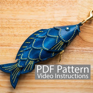 May include: A handcrafted, blue leather fish-shaped pouch with yellow stitching and a gold-colored zipper and clasp. The fish scales are detailed with yellow thread. The pouch is lying on a wooden surface. Text on the image reads "PDF Pattern Video Instructions."