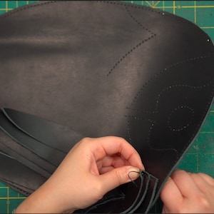 May include: A person is hand-stitching black leather strips to a larger piece of black leather. The larger piece has a decorative design stitched into it.