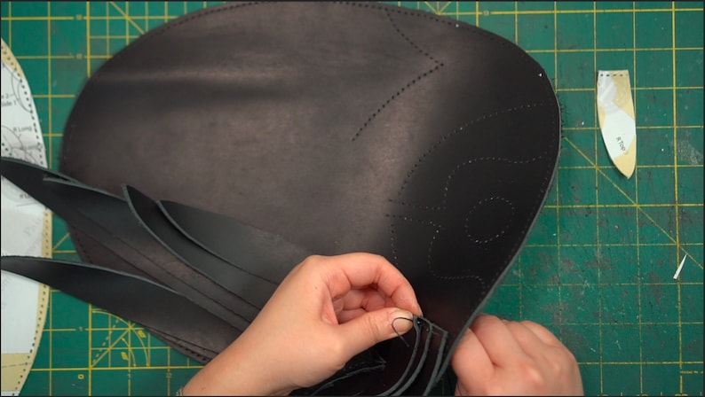 May include: A person is hand-stitching black leather strips to a larger piece of black leather. The larger piece has a decorative design stitched into it.