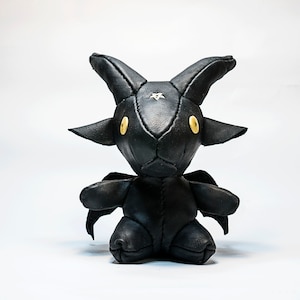 May include: A black leather stuffed toy shaped like a goat with horns and wings. The toy has yellow button eyes and a white star on its forehead.