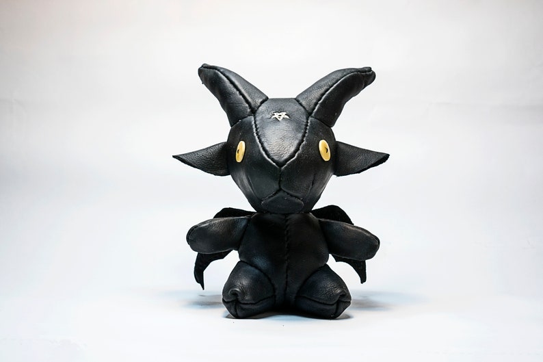 May include: A black leather stuffed toy shaped like a goat with horns and wings. The toy has yellow button eyes and a white star on its forehead.