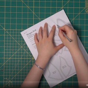 May include: A person is cutting out a pattern for a dragon doll. The pattern is printed on white paper and includes instructions for sewing the doll. The person is using a craft knife to cut out the pattern pieces.