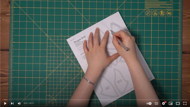 May include: A person is cutting out a pattern for a dragon doll. The pattern is printed on white paper and includes instructions for sewing the doll. The person is using a craft knife to cut out the pattern pieces.