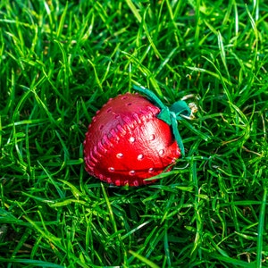 May include: A red leather strawberry keychain with green stitching and a green leather leaf. The keychain is lying on a bed of green grass.
