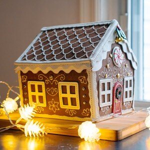 May include: A gingerbread house with brown roof tiles, white frosting trim, and decorative details. The house has a red door, white-framed windows, and a candy cane accent. It sits on a wooden board, illuminated by string lights.