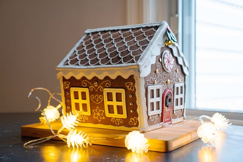 May include: A gingerbread house with brown roof tiles, white frosting trim, and decorative details. The house has a red door, white-framed windows, and a candy cane accent. It sits on a wooden board, illuminated by string lights.