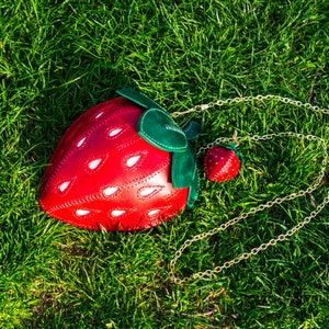 May include: A red leather strawberry-shaped handbag with a gold chain strap. The bag has white stitching and a green leather leaf detail.