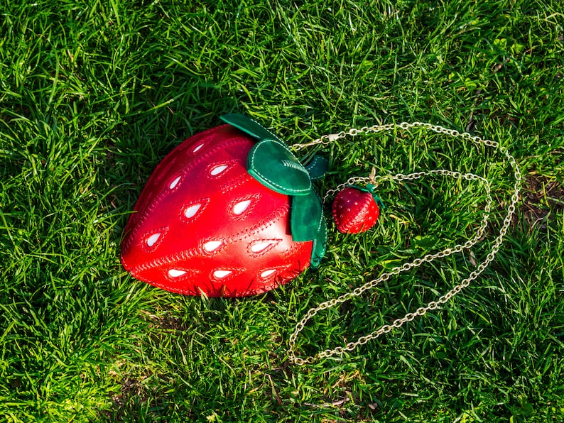 May include: A red leather strawberry-shaped handbag with a gold chain strap. The bag has white stitching and a green leather leaf detail.