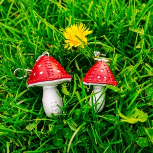 Peut inclure: Deux porte-cl&eacute;s en forme de champignon rouge et blanc avec des points blancs sur les chapeaux rouges. Les porte-cl&eacute;s sont pos&eacute;s sur un lit d'herbe verte.