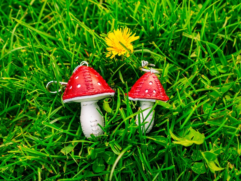 Peut inclure: Deux porte-cl&eacute;s en forme de champignon rouge et blanc avec des points blancs sur les chapeaux rouges. Les porte-cl&eacute;s sont pos&eacute;s sur un lit d'herbe verte.