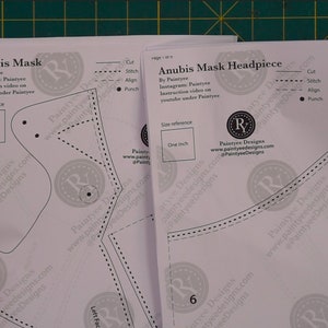 May include: A set of three white paper patterns for crafting an Anubis mask. The patterns include instructions for cutting, stitching, aligning, and punching. The patterns are labeled "Anubis Mask" and "Anubis Mask Headpiece".