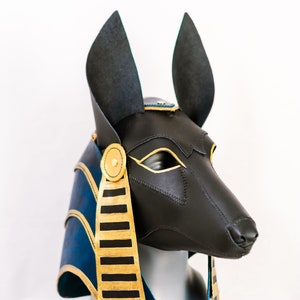 May include: A black leather Anubis mask with gold accents and blue and gold trim. The mask is on a mannequin head.