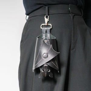 May include: Black leather bat-shaped accessory hanging from a silver clip attached to black pants. The bat has white stitching and a button detail. The accessory is a unique, gothic-inspired item.