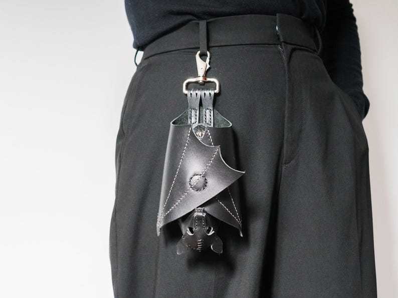 May include: Black leather bat-shaped accessory hanging from a silver clip attached to black pants. The bat has white stitching and a button detail. The accessory is a unique, gothic-inspired item.