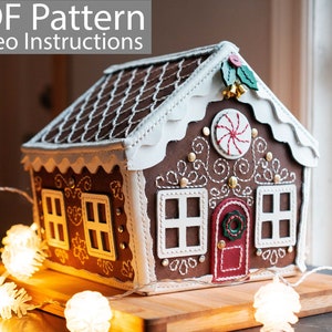 May include: A decorative gingerbread house with white trim, windows, and a red door. The roof is brown with white stitching. The house is decorated with a candy cane, holly, and small gold bells. The text "PDF Pattern Video Instructions" is at the top.
