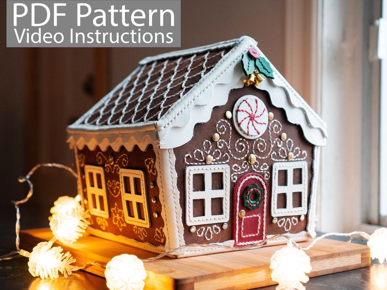 May include: A decorative gingerbread house with white trim, windows, and a red door. The roof is brown with white stitching. The house is decorated with a candy cane, holly, and small gold bells. The text "PDF Pattern Video Instructions" is at the top.